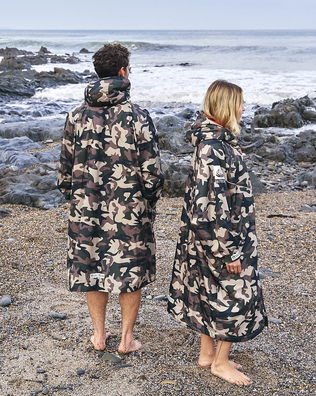 Two individuals standing on a rocky beach, wearing Saltrock's Recycled Four Seasons Changing Robe in Brown Camo, looking out at the sea.
