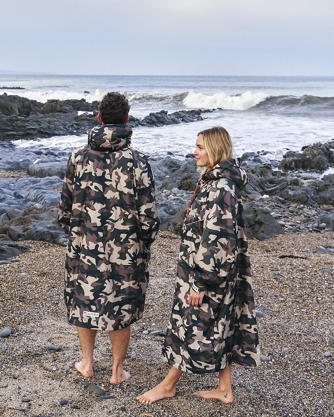 Two people wearing Saltrock's Recycled Four Seasons Changing Robe - Brown Camo standing on a beach looking at the sea.
