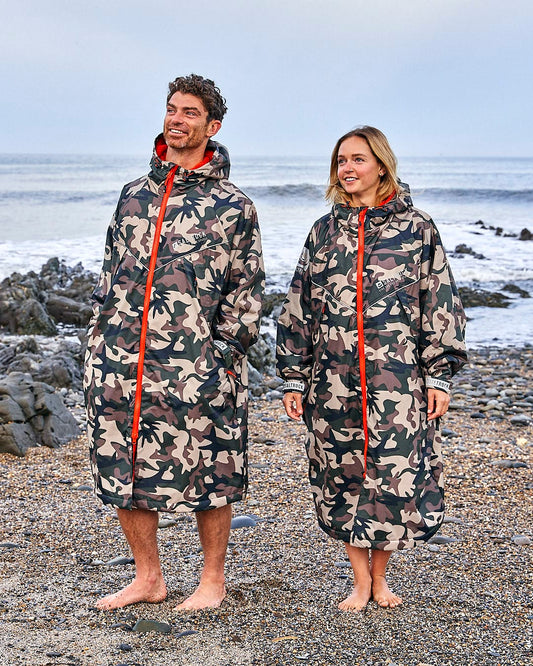 Two people in **Saltrock's Recycled Changing Robe - Brown Camo**, with red zippers, stand barefoot on a rocky beach, overlooking the ocean.