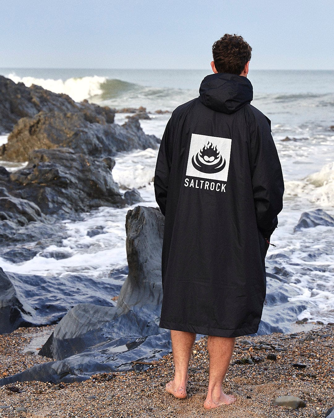 A man stands barefoot on a rocky beach facing the ocean, wearing a black hooded jacket with a "Saltrock" logo on the back. The jacket is actually a Recycled Four Seasons Changing Robe designed for beach.