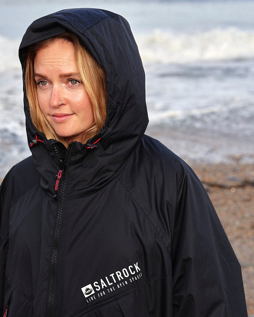 A woman standing on the beach wearing a Saltrock - Four Seasons - Waterproof Changing Robe - Black/Red.
