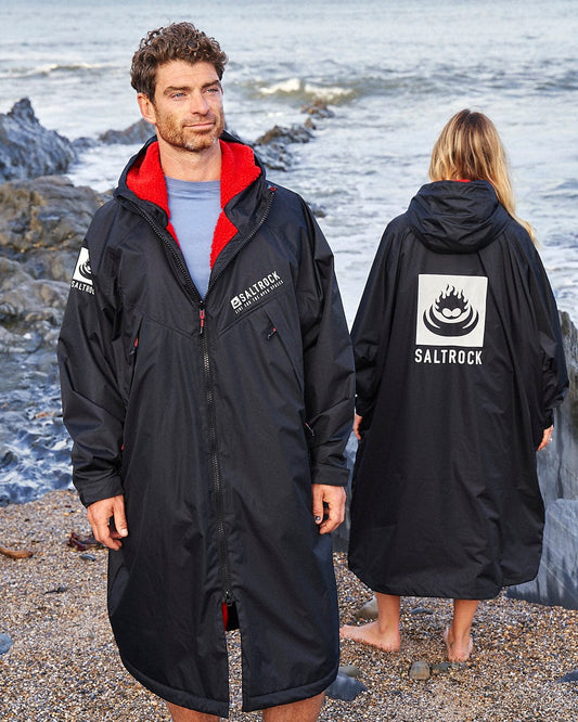 Two people stand on a rocky beach wearing black Saltrock Recycled Changing Robes - Black/Red, made from recycled materials. One faces forward, and the other turns away, both near the ocean. The Saltrock logo is visible on their robes as they embrace the coastal breeze.
