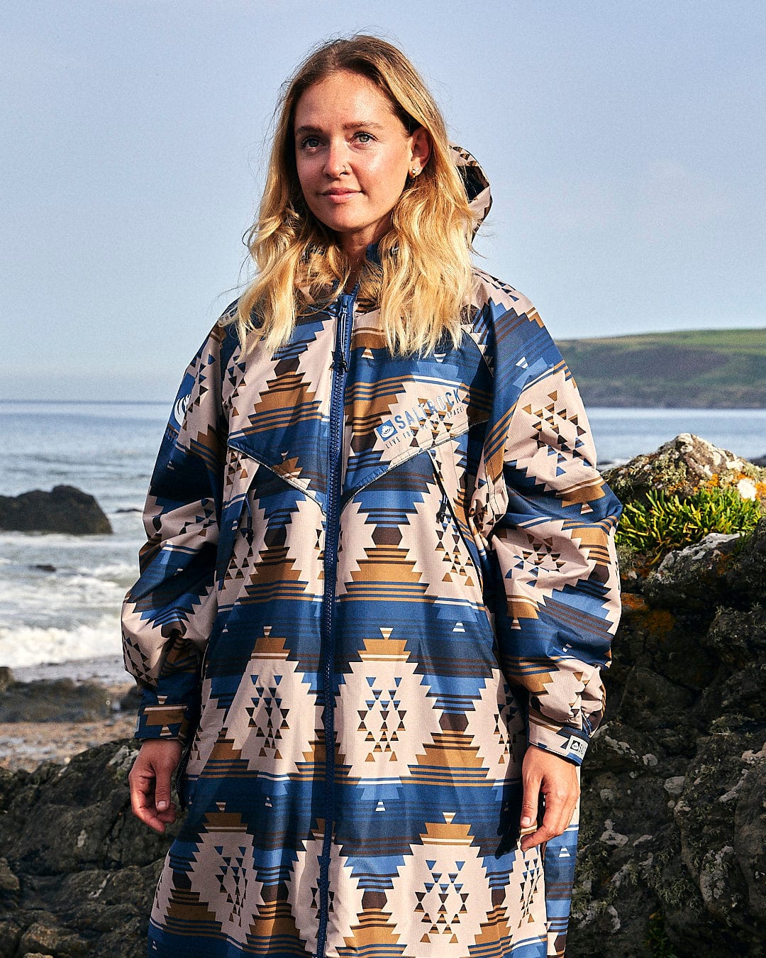 A woman wearing a Saltrock Four Seasons Changing Robe - Aztec print parka standing on rocks near the ocean.
