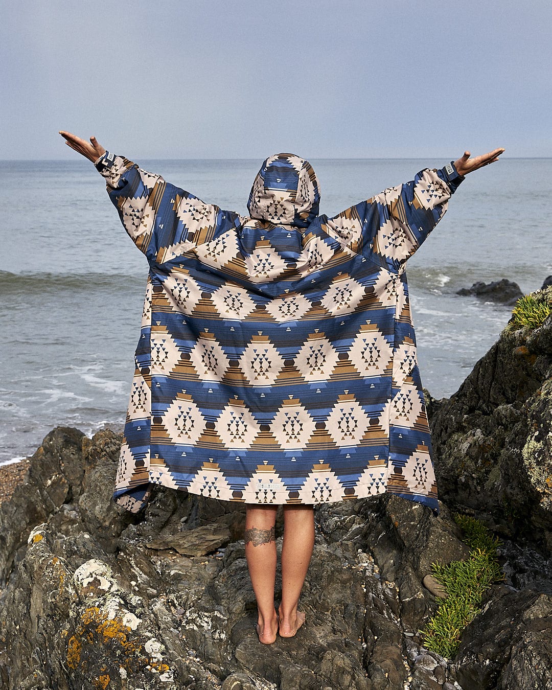 A woman standing on rocks with her arms outstretched wearing the Four Seasons Changing Robe - Aztec by Saltrock.