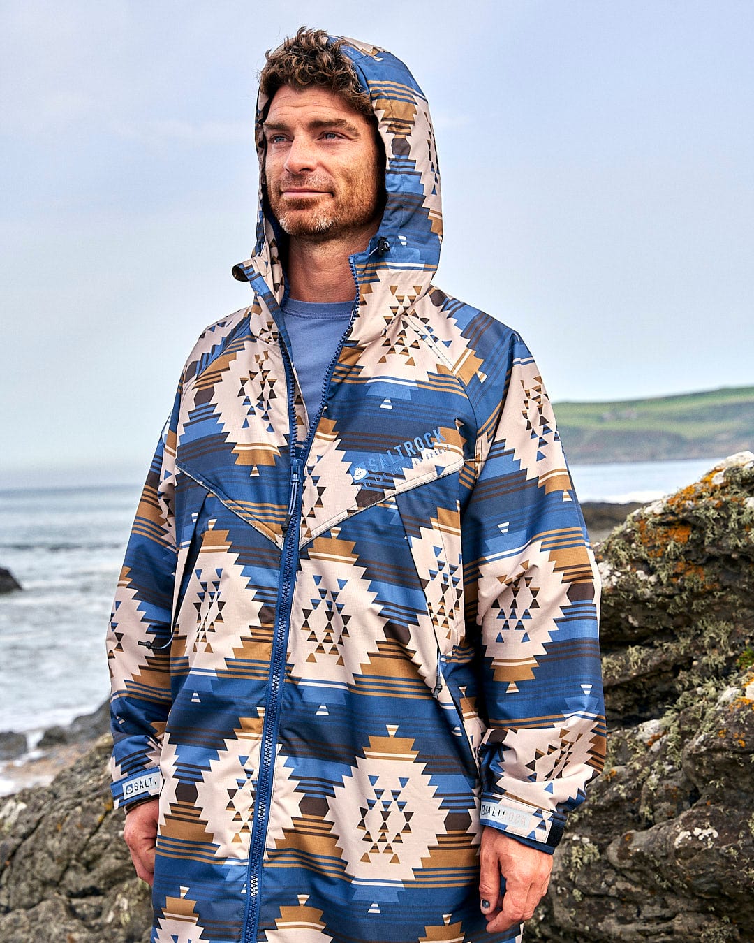 A man wearing a Saltrock Four Seasons Changing Robe - Aztec print raincoat standing on a rocky beach.