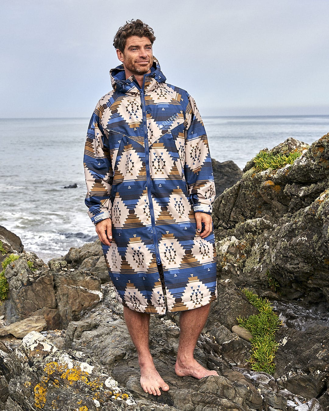 A man is standing on a rock next to the ocean wearing a Four Seasons Changing Robe - Aztec by Saltrock.