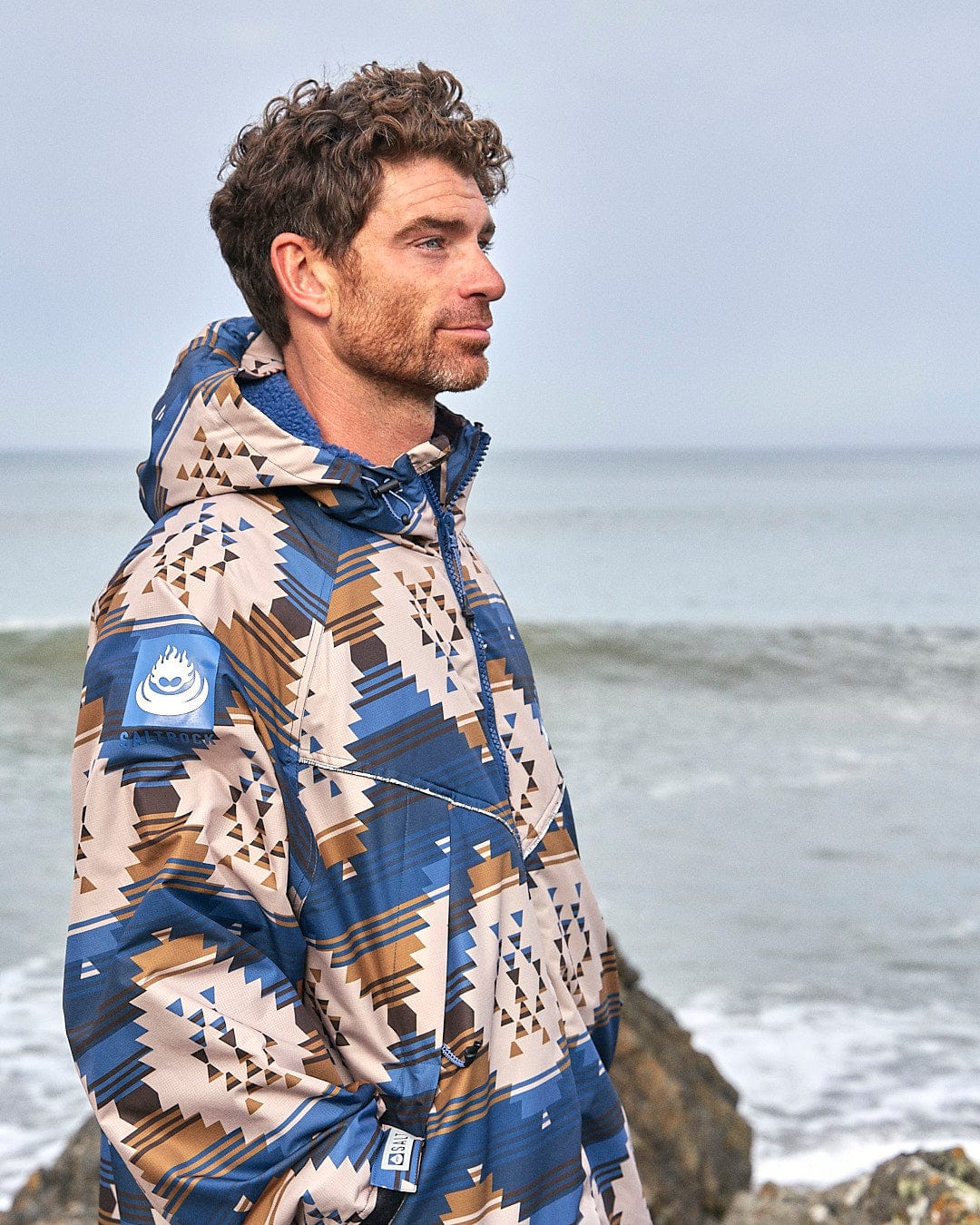 A man wearing a blue and brown Saltrock Four Seasons Changing Robe - Aztec print jacket standing on rocks near the ocean.