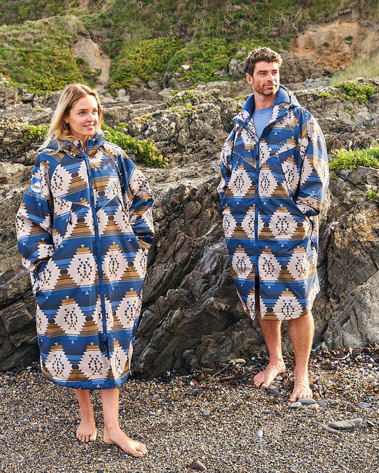 A man and woman standing on a rocky beach wearing the Saltrock Four Seasons Changing Robe - Aztec.