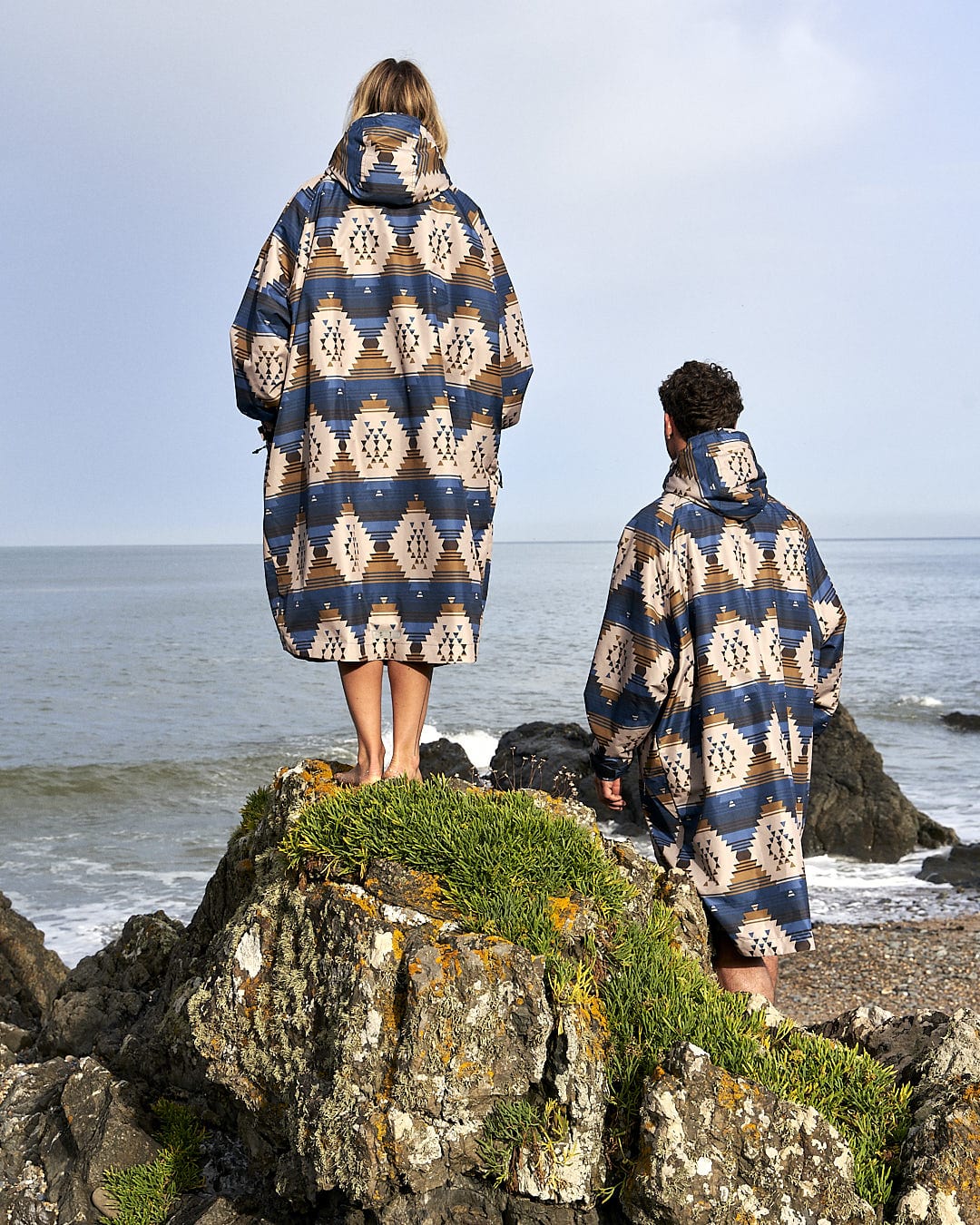 Two people standing on a rock next to the ocean wearing the Four Seasons Changing Robe - Aztec by Saltrock.