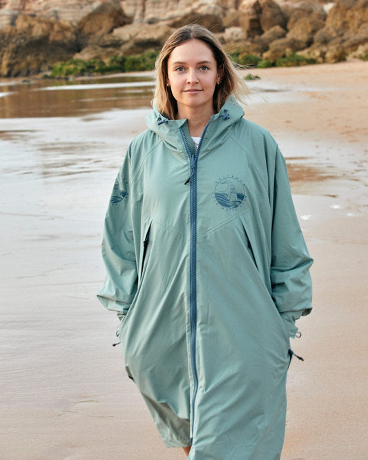 A person in a Saltrock Recycled Changing Robe - Light Green stands on a sandy beach with rocks in the background.