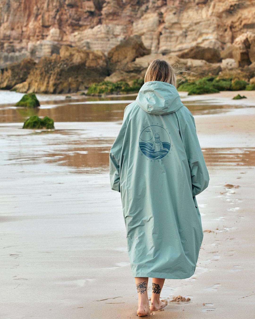 Person in a Saltrock Recycled Four Seasons Changing Robe - Light Green walking on a sandy beach toward rocky cliffs.