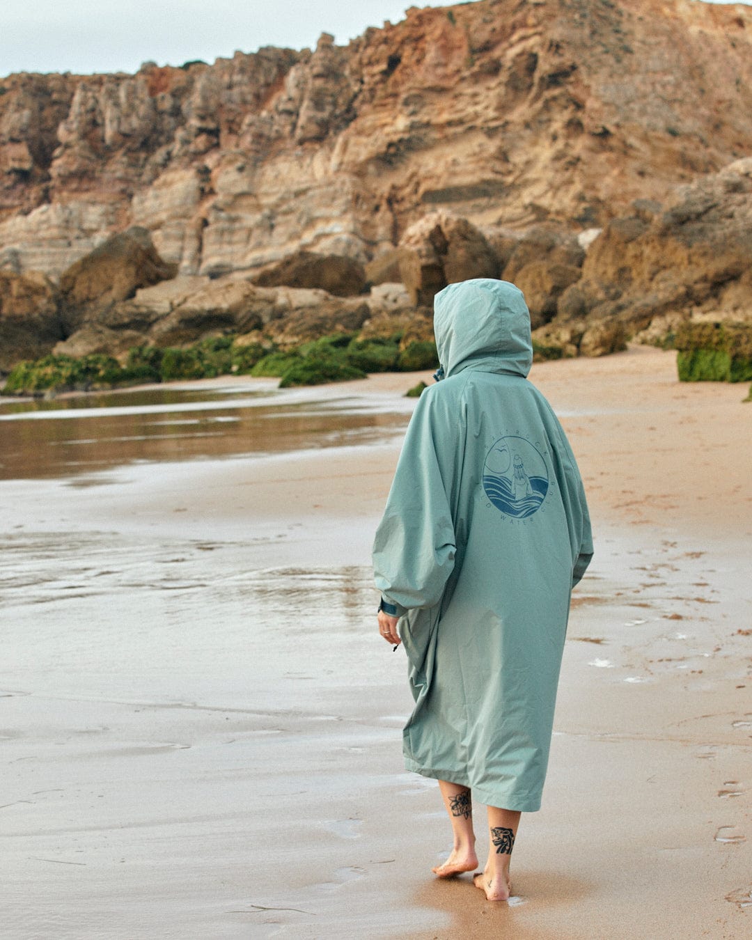 Person in a Saltrock Recycled Four Seasons Changing Robe in Light Green, walking along a sandy beach with cliffs in the background.