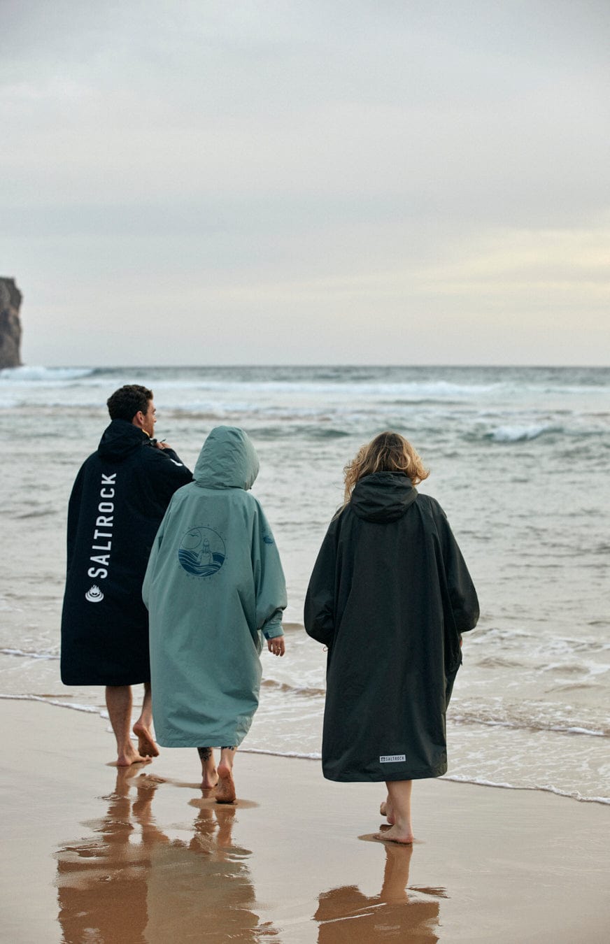 Three people wearing oversized Saltrock Recycled Changing Robes - Light Green made from recycled materials walk along a sandy beach with their backs to the camera, facing the ocean.