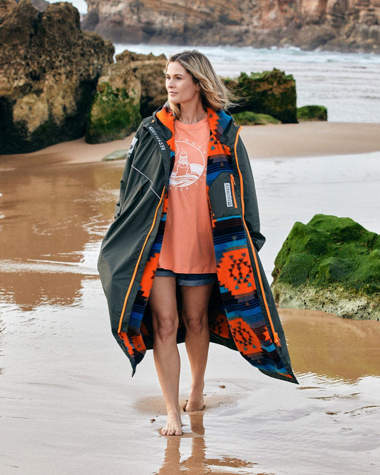 A woman in a black and colorful patterned coat walks barefoot on a beach with green rocks in the background. She is wearing an orange shirt and denim shorts, her Saltrock Recycled Changing Robe - Green/Aztec keeping her warm against the sea breeze.
