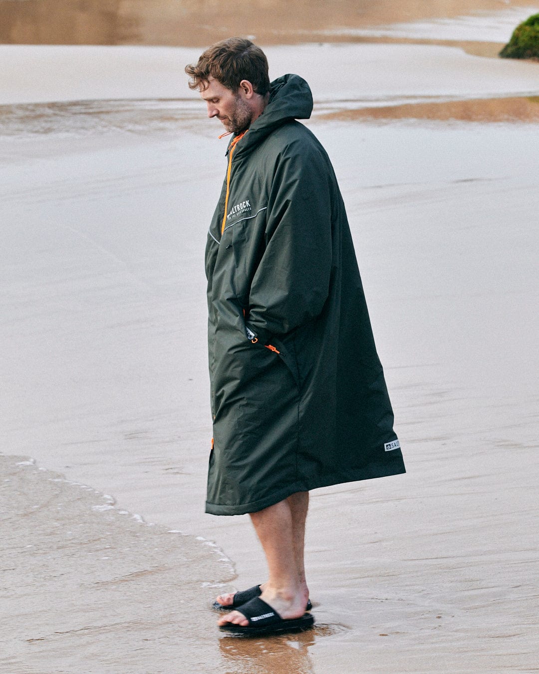 Man standing on wet sand wearing a Saltrock Recycled Four Seasons Changing Robe in Green/Aztec and sandals.