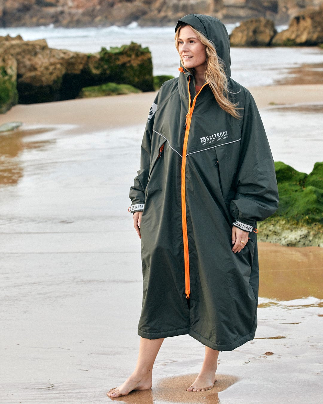 Woman standing on the beach wearing a long waterproof Recycled Four Seasons Changing Robe with fleece lining and a hood, made from recycled material by Saltrock.