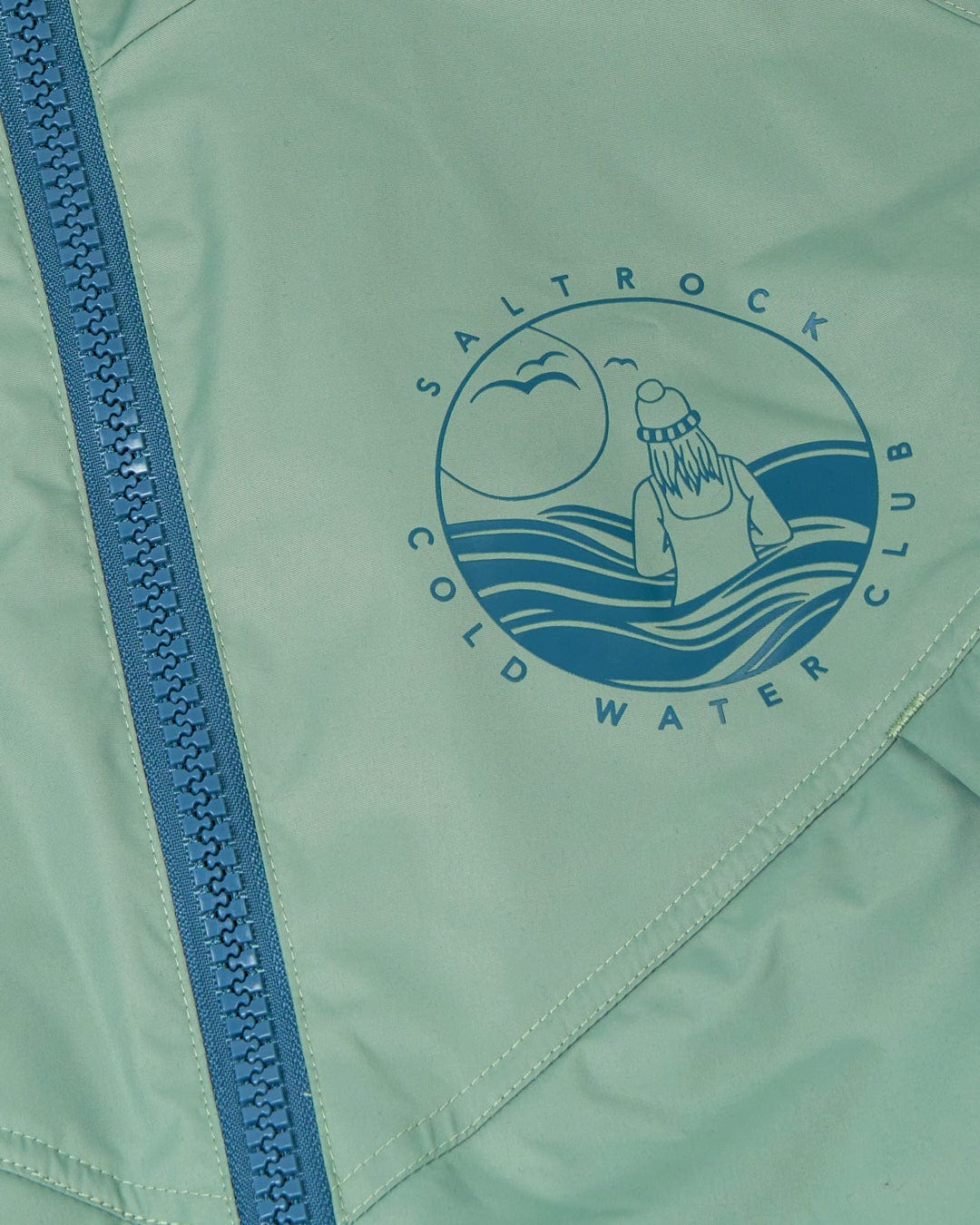 Close-up of a Saltrock Recycled Changing Robe - Light Green, showing a blue zipper and a circular logo with a person facing the sea, mountains, and sun. It comes with an adjustable hood and is part of the "Saltrock Cold Water Club.