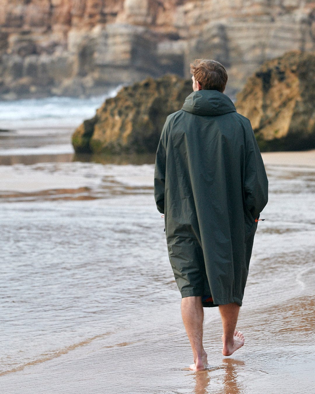 A person walking alone along a sandy beach with waves gently breaking at the shore, wearing a Saltrock jacket with fleece lining.