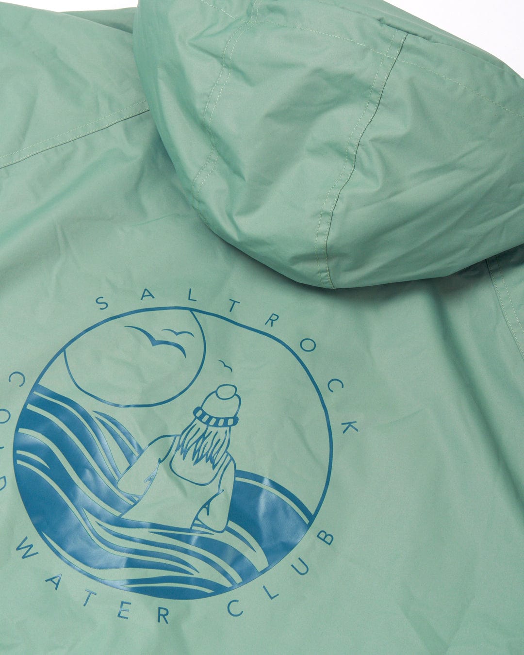 Close-up of the back of a green hooded jacket featuring a circular logo with an illustration of a person wearing a beanie watching waves and a seagull, along with the text "Saltrock Cold Water Club." This stylish piece, made from recycled materials, also features an adjustable hood for added comfort. Introducing the Recycled Changing Robe - Light Green by Saltrock.