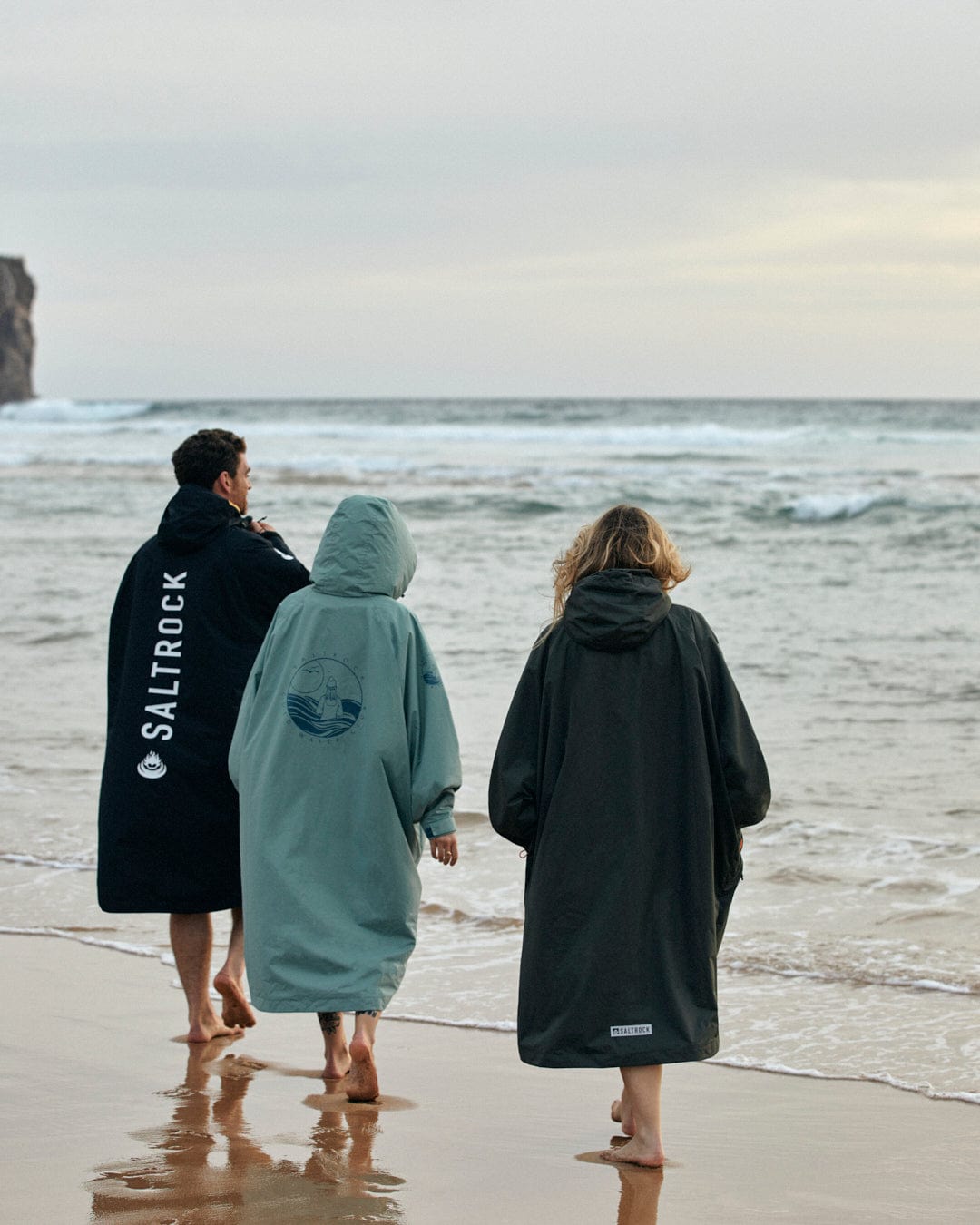 Three individuals walking on the beach wearing cozy Saltrock Recycled Four Seasons Changing Robes in Green/Aztec with fleece lining.