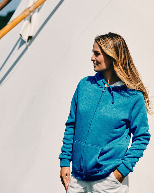 A person with long hair wearing a blue Saltrock Galak Women's Fur Lined Hoodie and white pants stands outdoors, looking to their right, ready for an adventure.