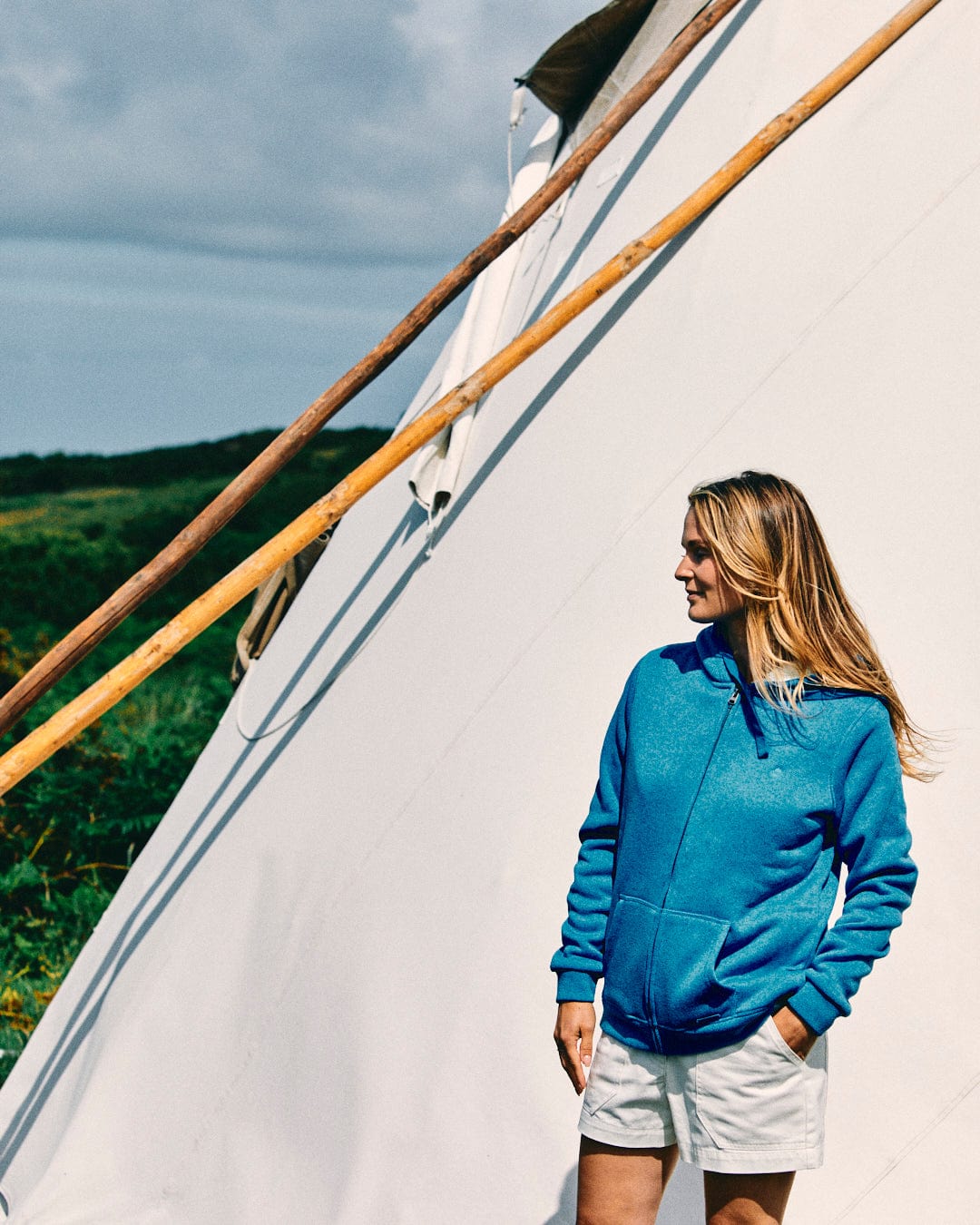 A person wearing the Saltrock Galak Women's Fur Lined Hoodie in blue and beige shorts stands in front of a white tent, looking to the side. Green hills and a cloudy sky can be seen in the background, hinting at an adventure waiting to unfold.
