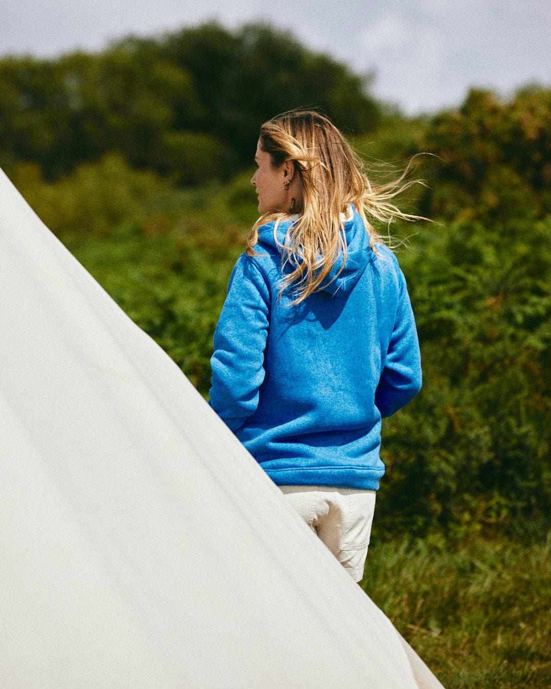 A person wearing the Galak - Womens Fur Lined Hoodie in blue by Saltrock stands outdoors facing away, with greenery and a white tent in the foreground.