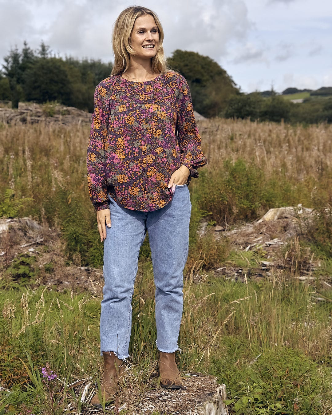 A woman standing in a field wearing Garnet - All Over Print Blouse - Orange by Saltrock jeans and a floral top.