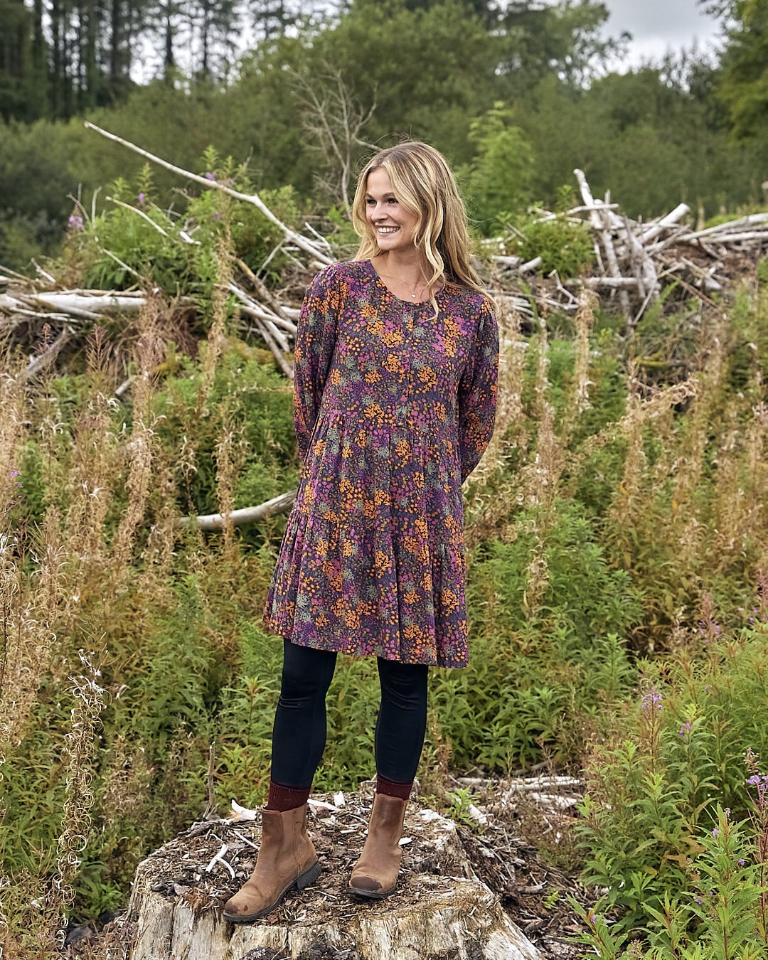 A woman in a Garnet - Womens Short Dress - Orange by Saltrock, standing on a stump in a field.