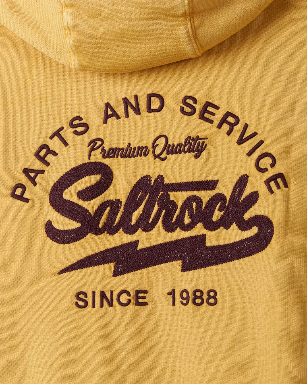 Close-up of a yellow Saltrock Gas Station hoodie with embroidered text "parts and service premium quality recycled polyester since 1988" in brown and purple thread.