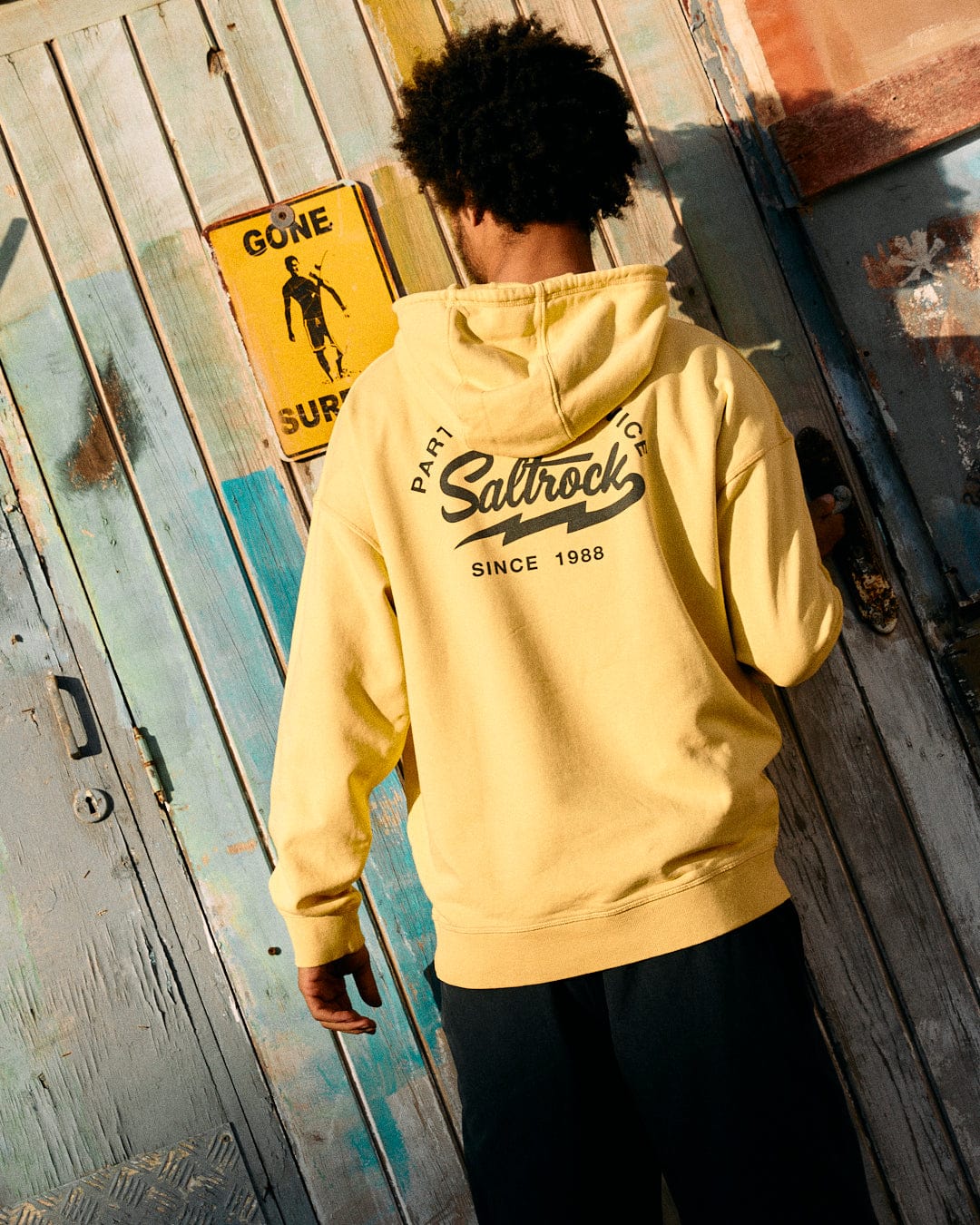 A person in a yellow Saltrock Gas Station - Recycled Mens Pop Hoodie stands in front of a colorful wooden wall with a "gone surfing" sign.