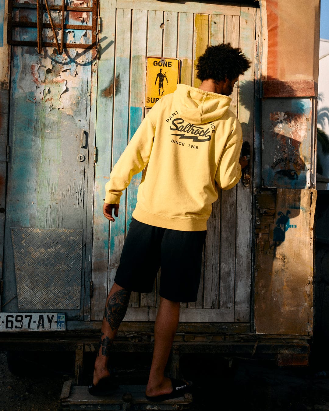 A person in a yellow Saltrock Gas Station - Recycled Mens Pop Hoodie and black shorts stands facing a colorful, weathered shipping container, with the text "gone surfing" visible above their head.