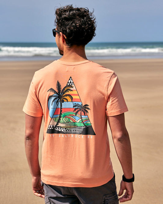 A man is standing on the beach wearing a Saltrock Geo Beach - Mens Short Sleeve T-Shirt - Coral.