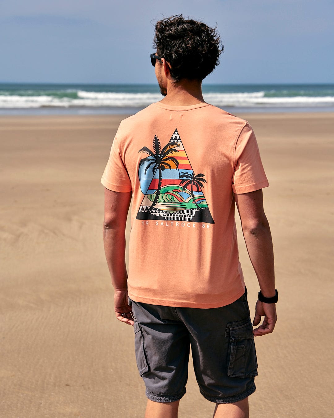 A man walking on the beach wearing a Saltrock Geo Beach - Mens Short Sleeve T-Shirt - Coral with a palm tree on it.