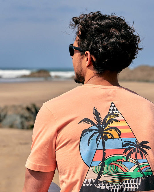 A man is standing on the beach wearing a Saltrock Geo Beach - Mens Short Sleeve T-Shirt - Coral with a palm tree on it.