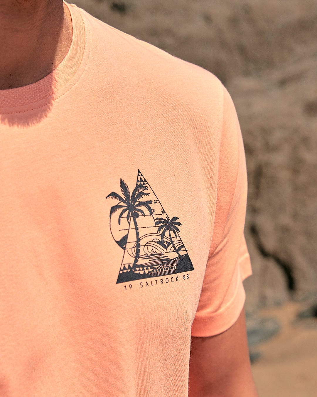 A man wearing a Saltrock Geo Beach - Mens Short Sleeve T-Shirt - Coral with a palm tree on it.