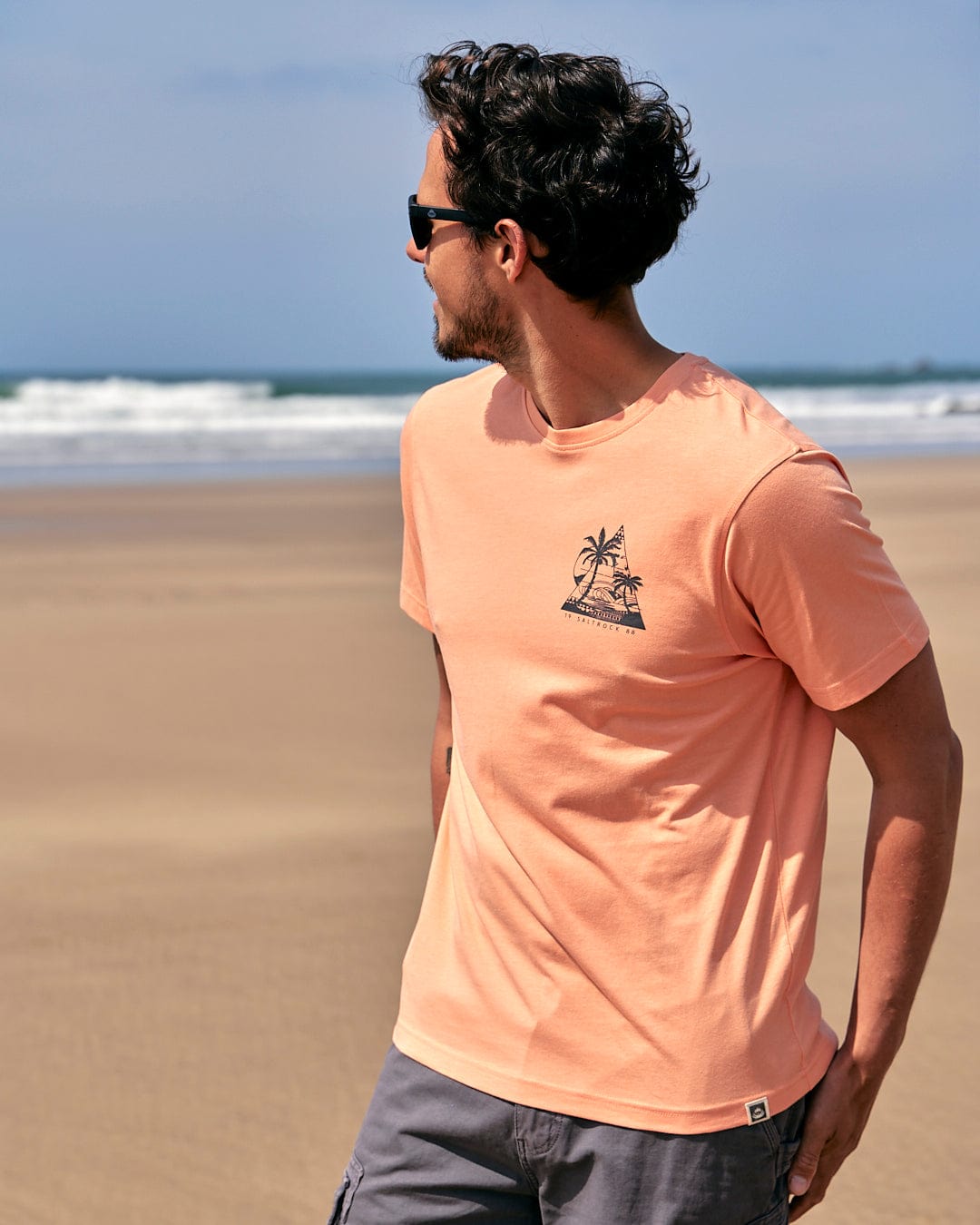 A man wearing a Saltrock Geo Beach - Mens Short Sleeve T-Shirt - Coral standing on the beach.