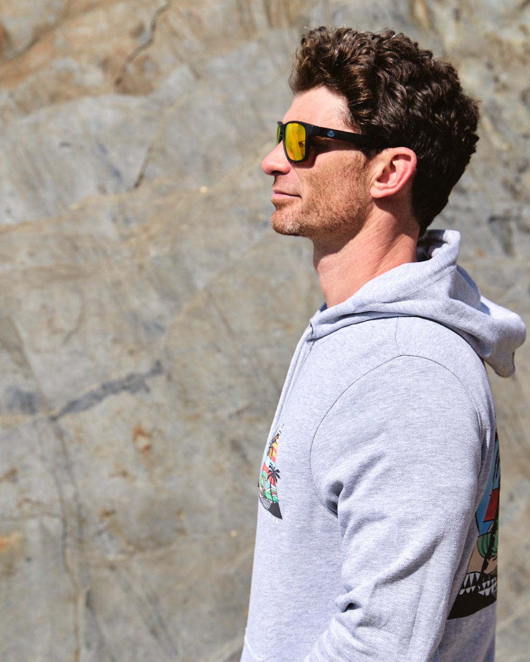 A man wearing "Saltrock - Geo Beach - Mens Pop Hoodie - Grey" sunglasses and a hoodie in front of a rock.