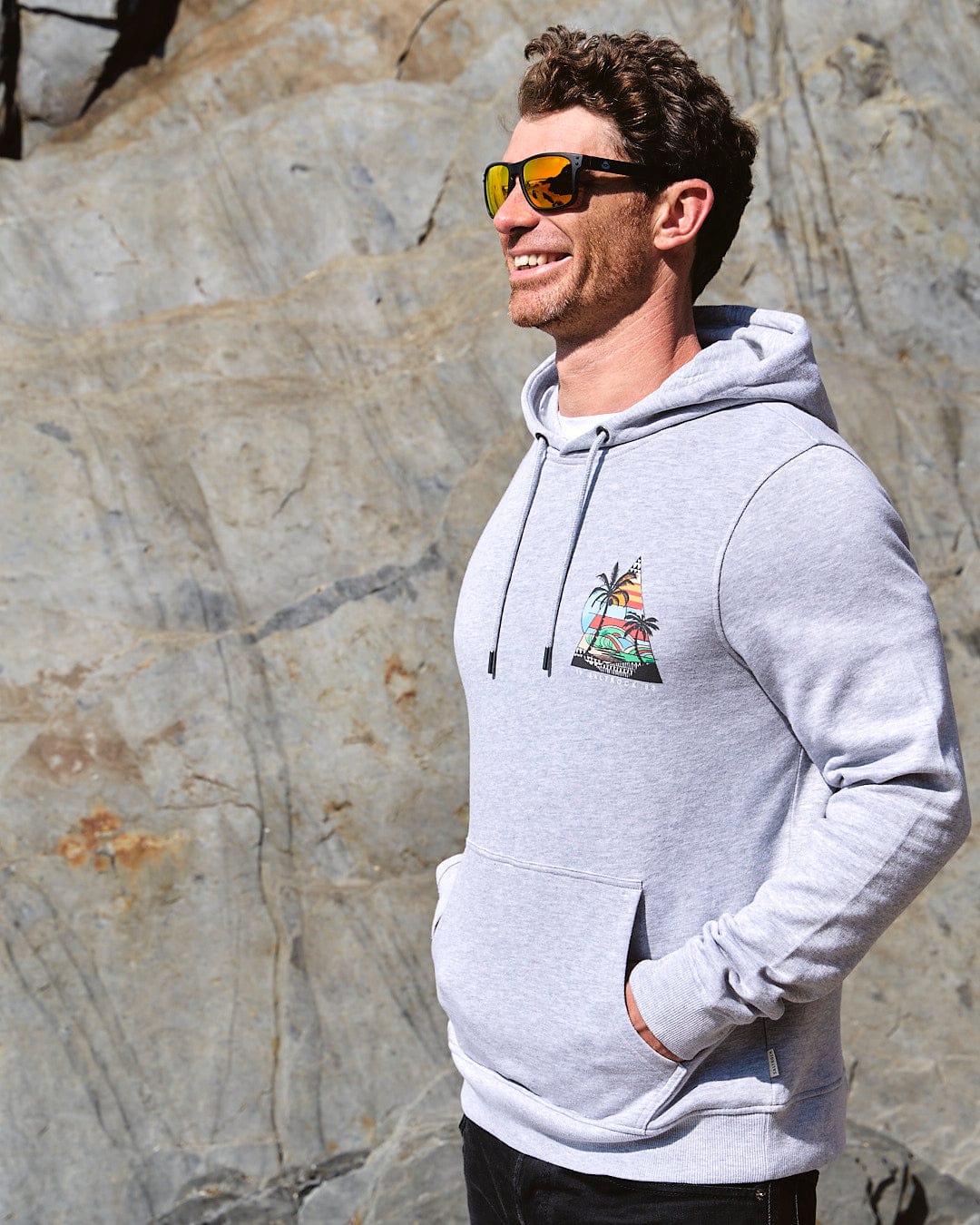 A man wearing sunglasses and a hoodie standing in front of rocks.