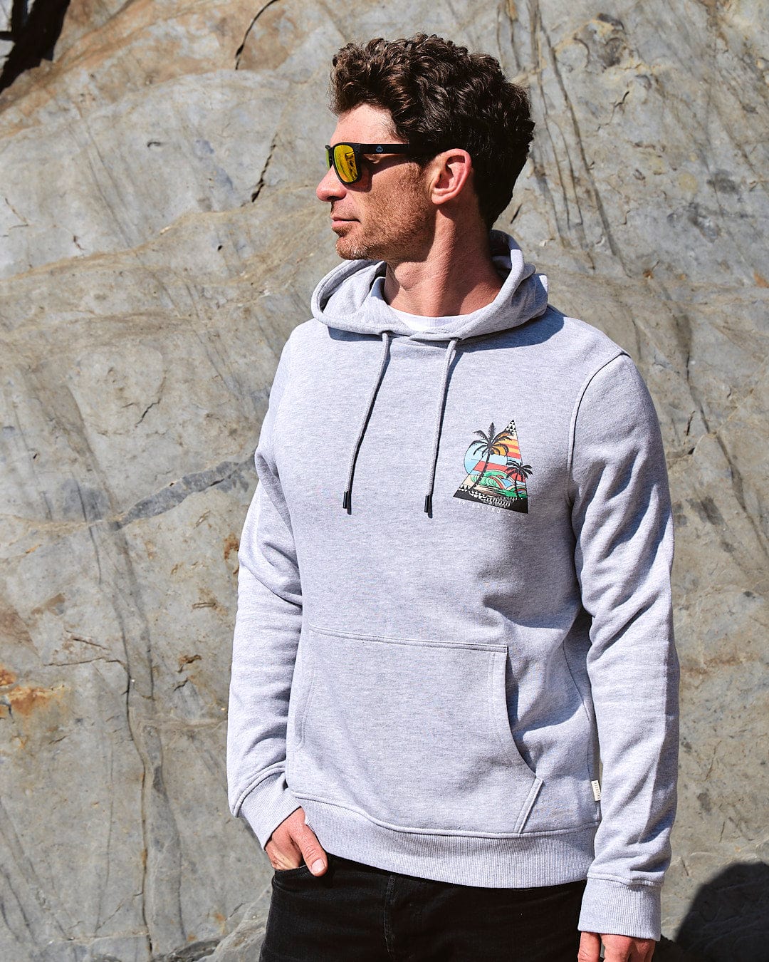 A man wearing sunglasses and a Geo Beach - Mens Pop Hoodie - Grey by Saltrock standing in front of a rock.
