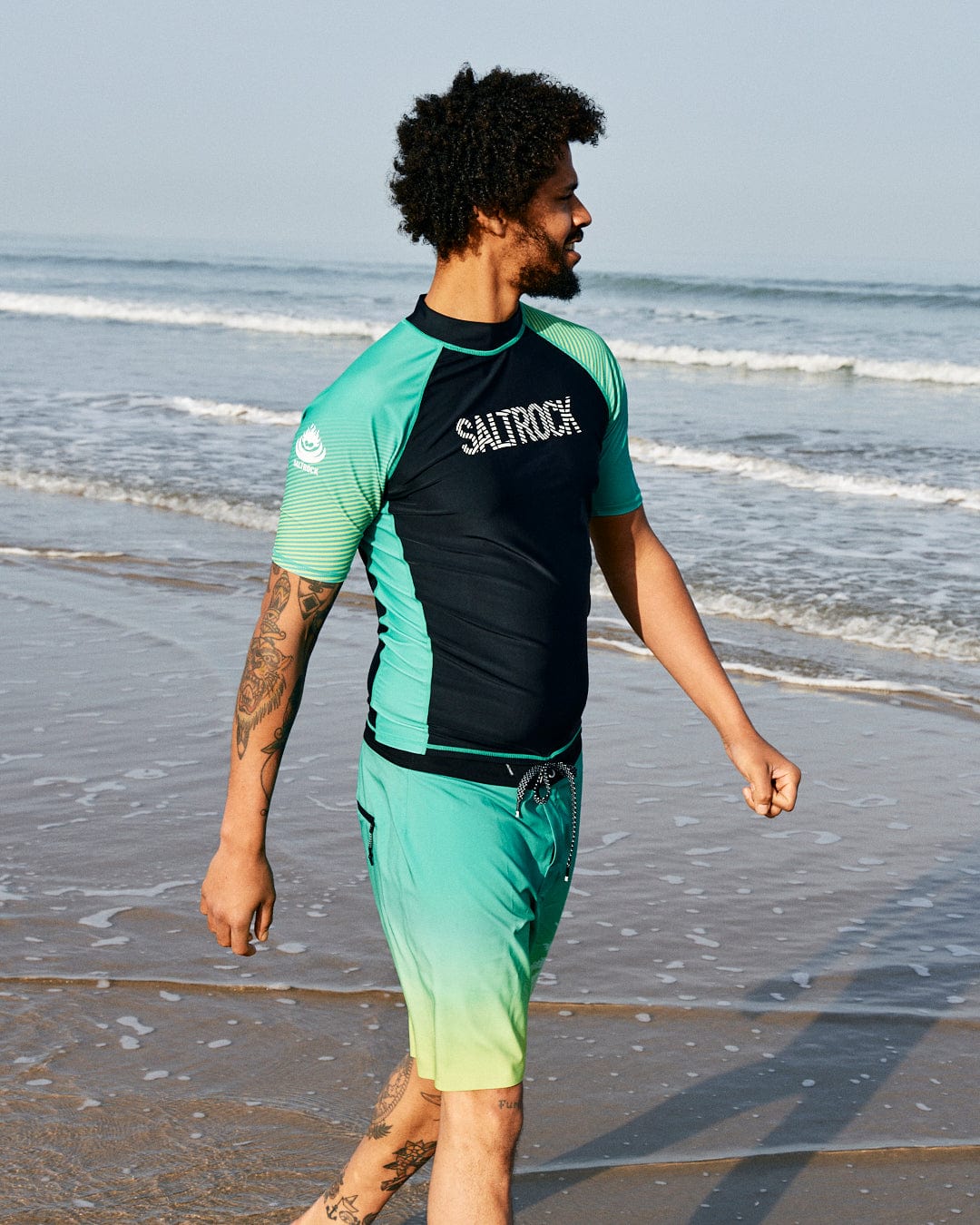 A man with curly hair wearing a rash guard and Saltrock Geo Palms Men's Boardshorts in Green, made from Repreve recycled material, walking on the beach and looking to the side.