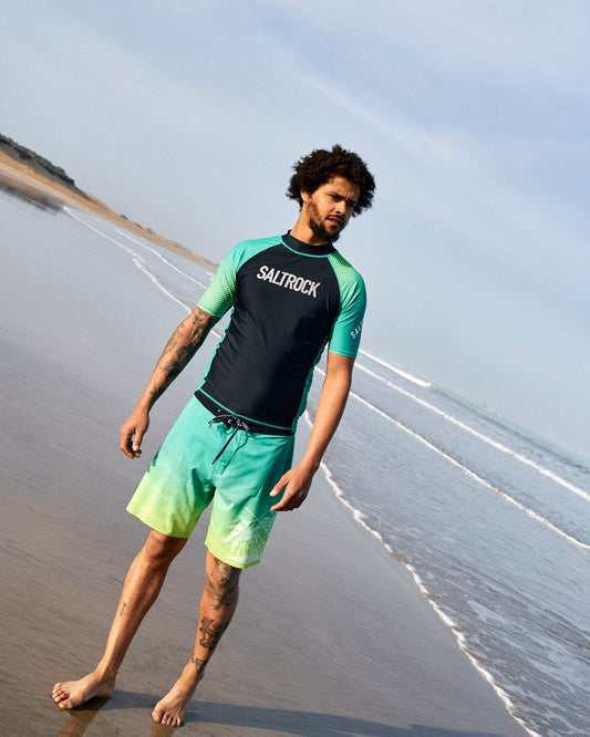 A man wearing a teal Saltrock t-shirt with ombre palm print and Saltrock Geo Palms boardshorts in green stands barefoot on a sandy beach, looking thoughtfully to the side.