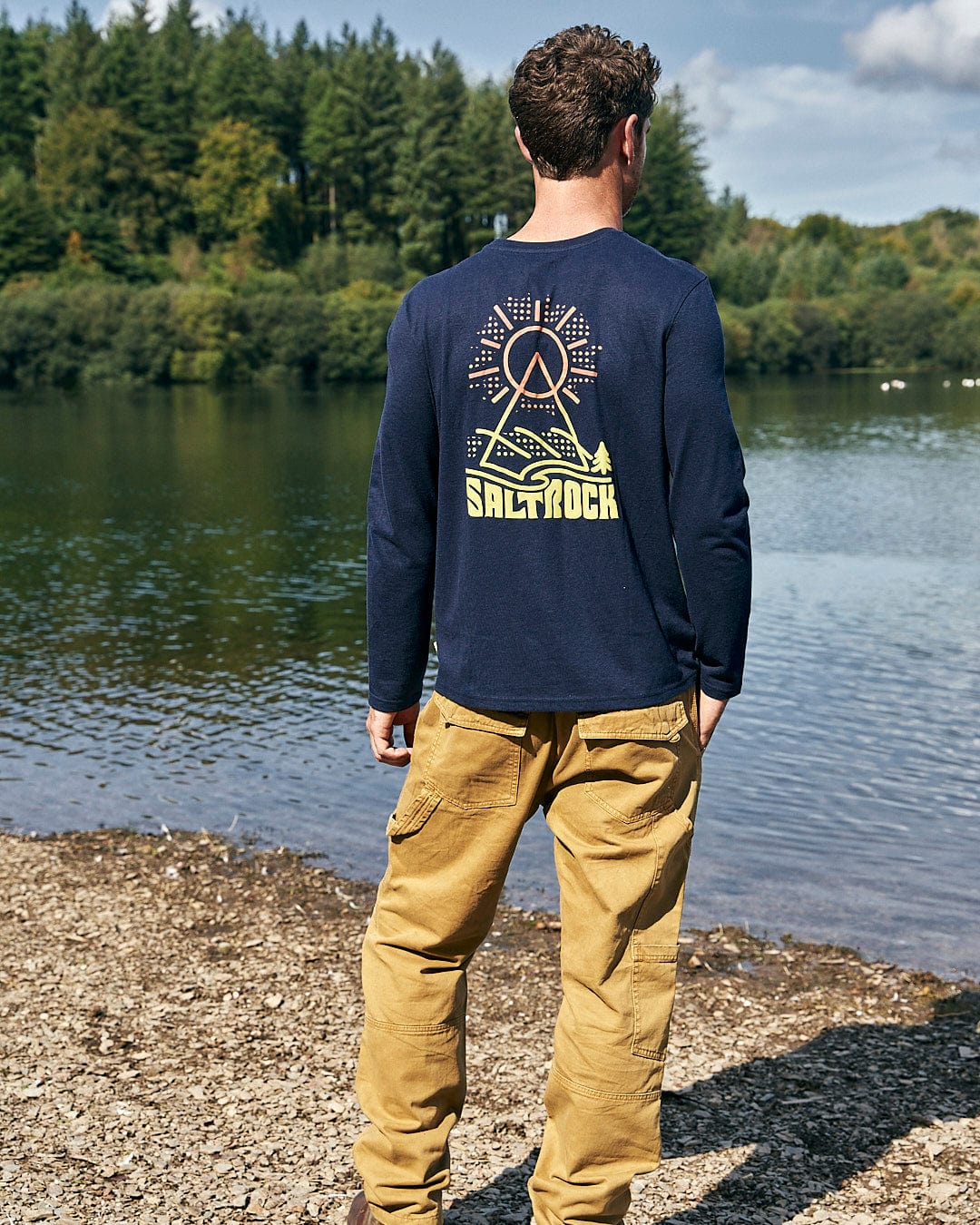 A man standing by a lake wearing a Saltrock Geo Peak - Mens Long Sleeve T-Shirt - Dark Blue.