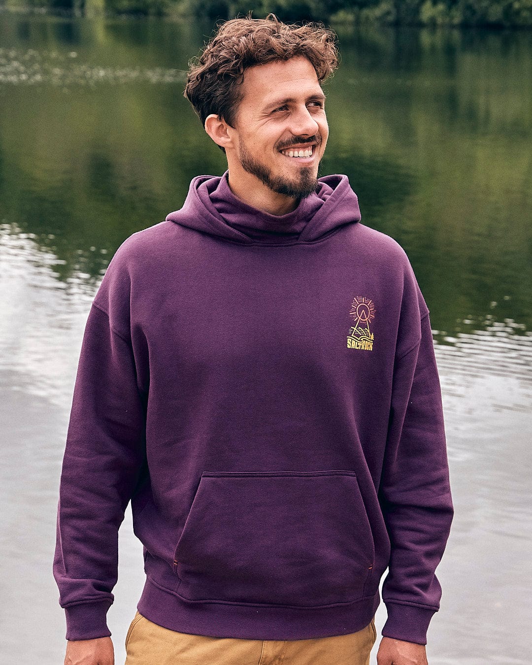 A man wearing a Saltrock Geo Peak - Mens Oversized Snood Pop Hoodie - Dark Purple standing by a lake.
