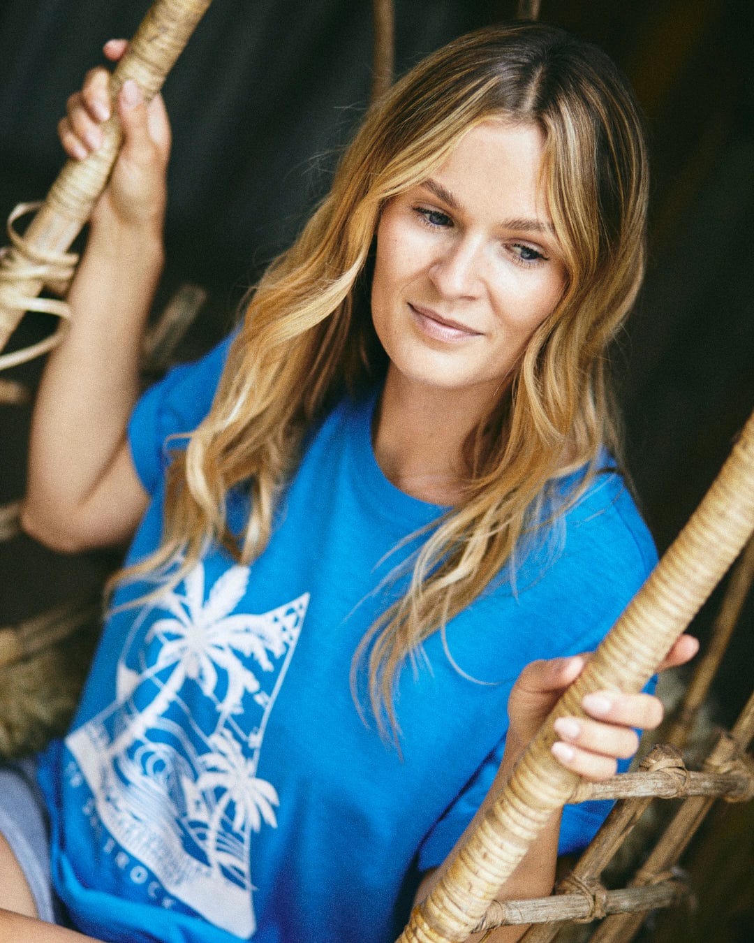 A woman with long blonde hair sits on a woven swing chair, wearing the Saltrock Geo Beach Mono Women's T-Shirt in blue, which features a palm tree design and is made of 100% cotton, perfectly showcasing its relaxed vibe.