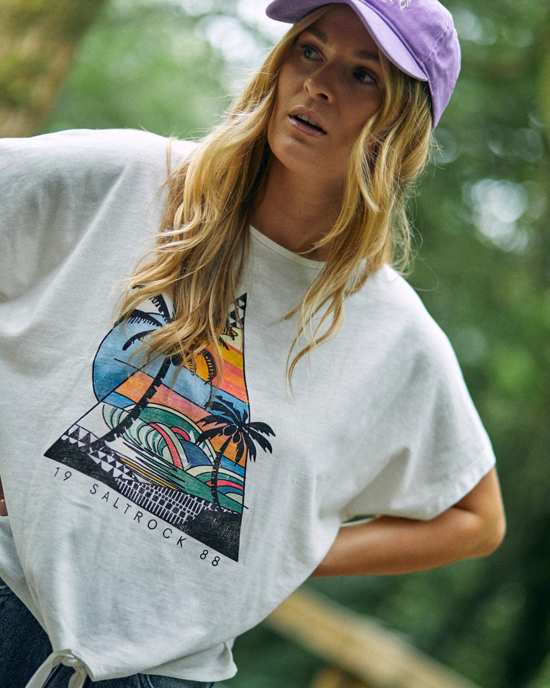 A person with long hair wearing a purple cap and Saltrock's Geo Beach Women's Tie Front T-Shirt in white, featuring a colorful graphic design, stands outdoors.