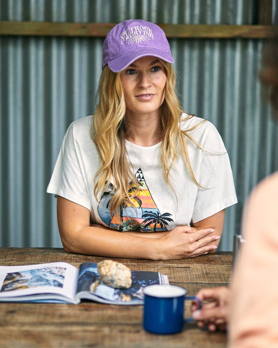 Woman wearing a purple cap and the Saltrock Geo Beach Women's Tie Front T-Shirt in white, sitting at a wooden table with a magazine, rock, and blue coffee mug in front of her. Corrugated metal wall in the background.