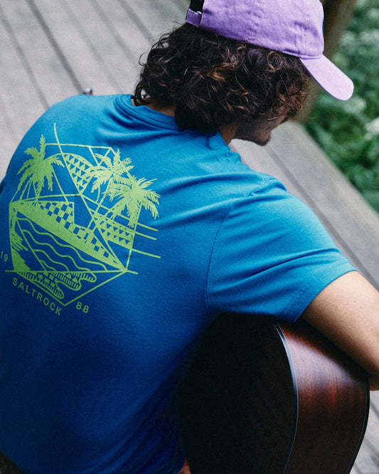A person wearing the Saltrock Geo Palms Solid Men's Recycled Hybrid T-Shirt in blue, featuring a beach and palm tree design, sits on a wooden deck while holding a guitar and sporting a purple cap.