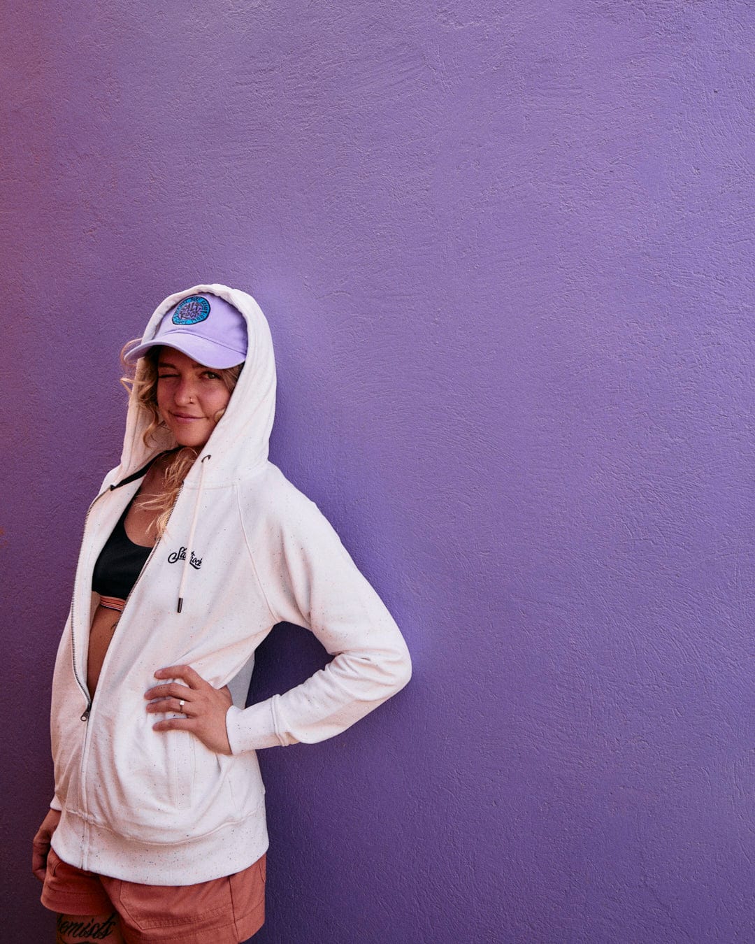 Woman in a Ginny - Womens Zip Hoodie in Cream by Saltrock and pink shorts crafted from peached soft hand feel fabric, standing against a purple wall, smiling with hood over her head.