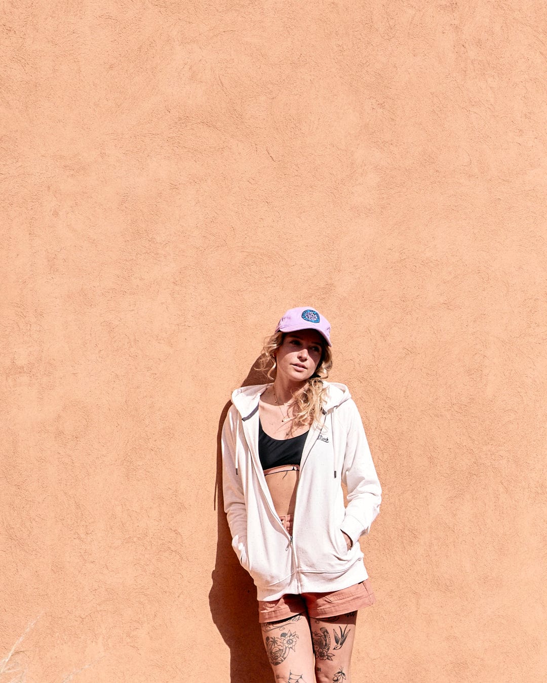 A woman in a Ginny - Womens Zip Hoodie - Cream from Saltrock, standing against an orange wall, looking to the side with a relaxed pose.